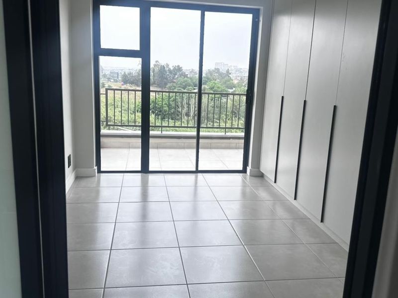 A bright, modern bedroom featuring neutral tiled flooring, ample built-in cupboard space, and a sliding glass door leading to a private balcony.