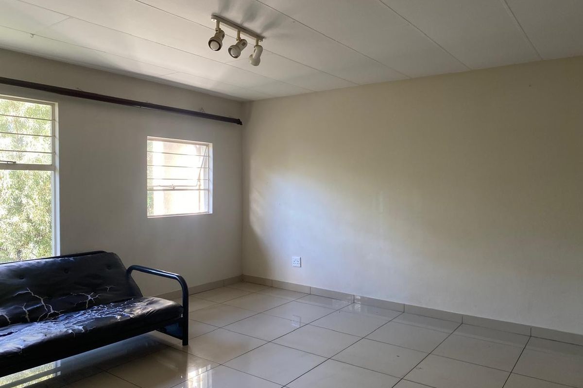 A sparsely furnished living area featuring tiled flooring, track lighting, and casement windows with security bars.