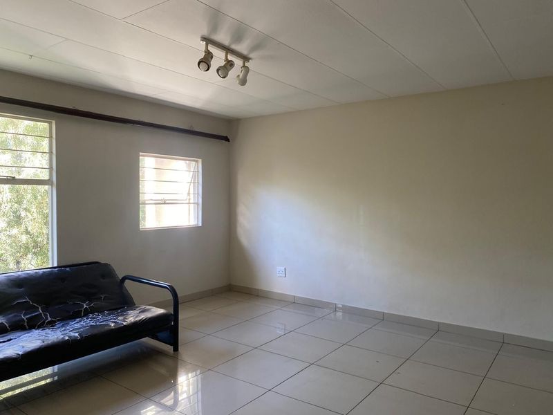 A sparsely furnished living area featuring tiled flooring, track lighting, and casement windows with security bars.