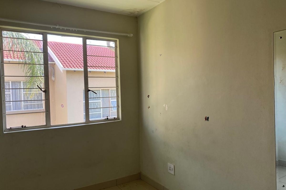 An empty bedroom featuring tiled flooring, security bars on the window, and a wall-mounted mirror, requiring cosmetic updates to the walls and ceiling.
