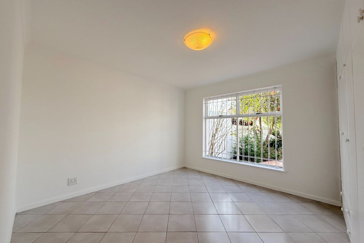 A bright, empty bedroom featuring neutral tiled flooring, built-in cupboards, and a window fitted with security bars.