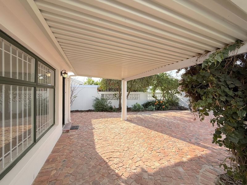 A covered outdoor patio area featuring brick paving, a corrugated roof structure, and a view of the enclosed garden and perimeter wall.