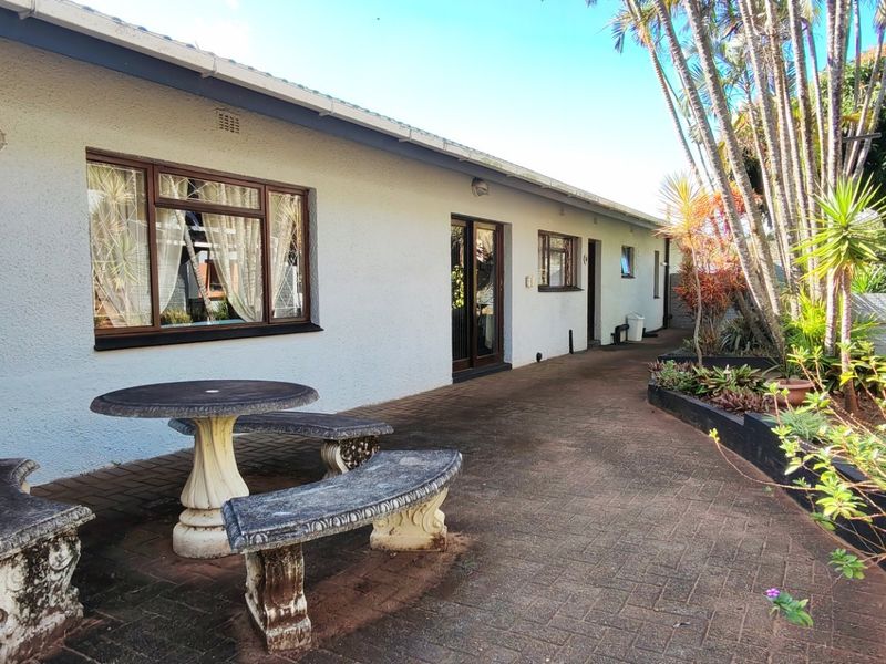 A rear exterior view of the house featuring a paved patio area, established garden beds, and access via sliding doors.
