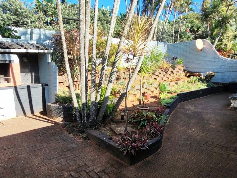 A covered outdoor patio area featuring a built-in braai, brick paving, and a landscaped garden bed with retaining walls.