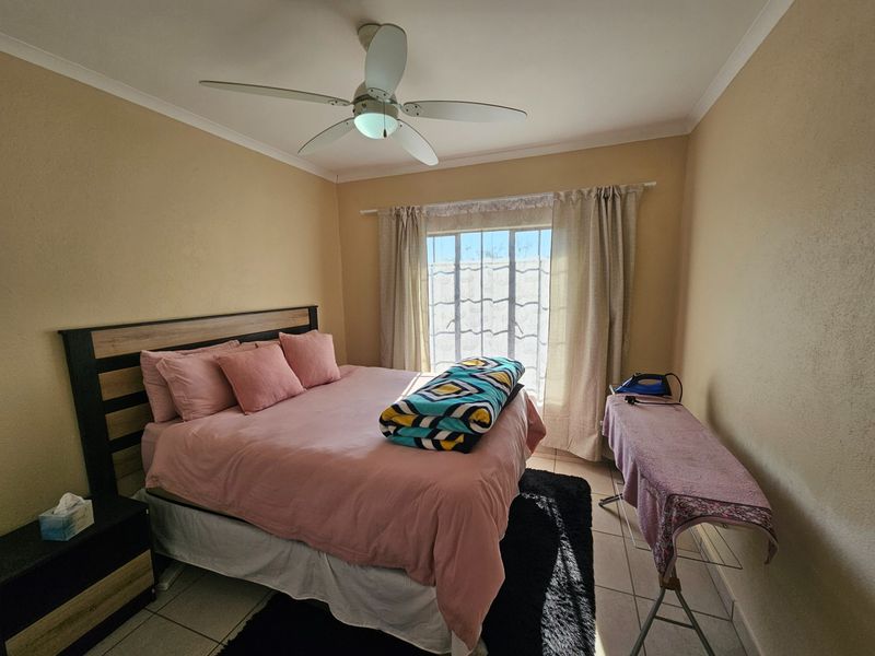A functional bedroom featuring tiled flooring, a ceiling fan, and a window with security bars, providing a comfortable and practical living space.
