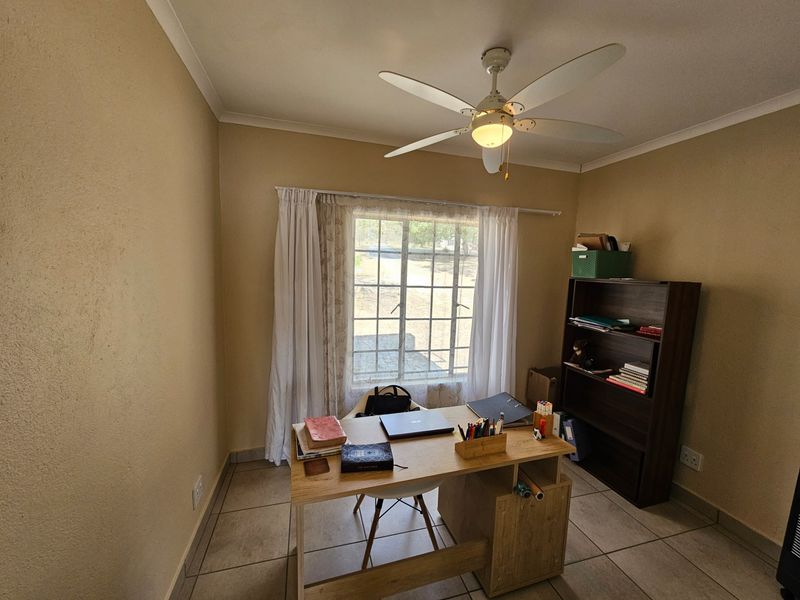 A functional home office space featuring tiled flooring, a ceiling fan, and a window with security bars, providing a practical workspace.