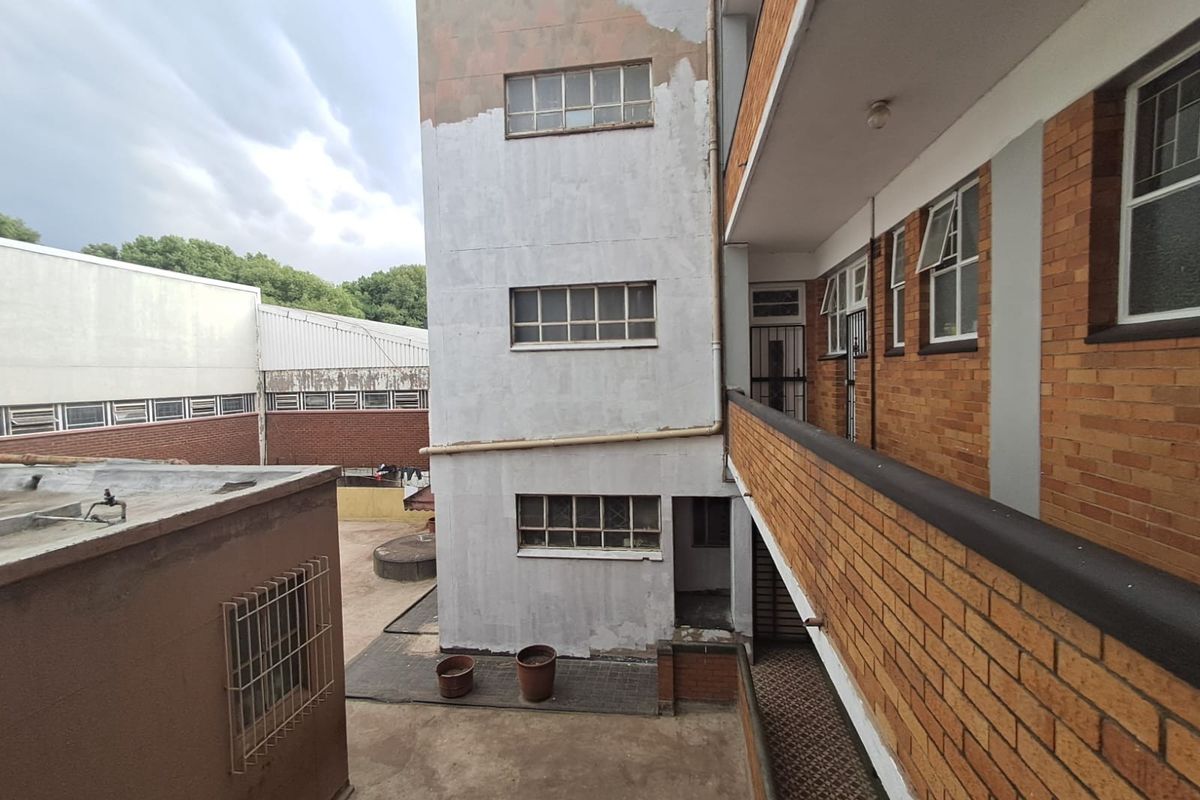 Rear exterior view of an apartment building featuring brick facade, steel-framed windows, and a concrete walkway with visible signs of weathering and paint deterioration.