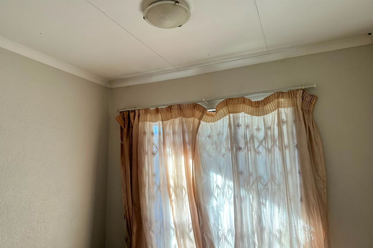A bedroom interior featuring a ceiling light fixture, curtain rail, and security gate over the window, showing signs of wear on the walls and ceiling.