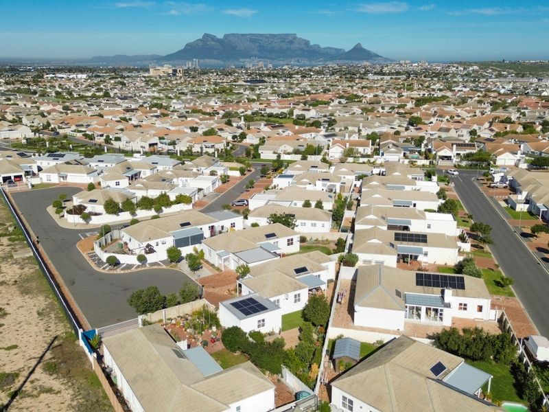 An aerial view of a residential neighborhood featuring numerous homes with tiled roofs, solar panels, and clear views of Table Mountain in the distance.