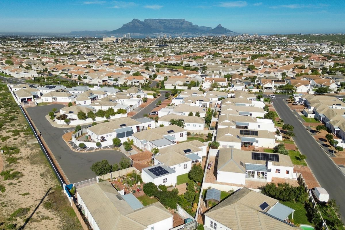 An aerial view of a residential neighborhood featuring numerous homes with tiled roofs, solar panels, and clear views of Table Mountain in the distance.