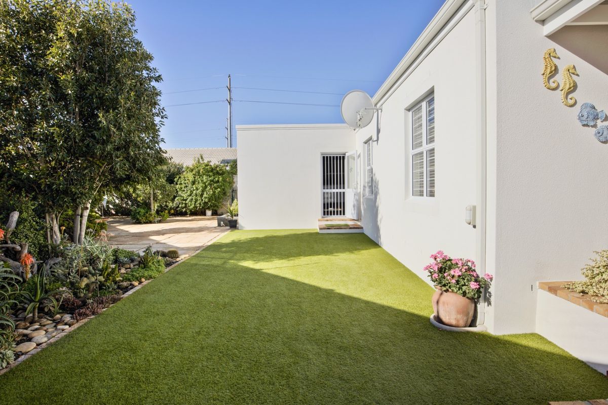 A well-maintained rear garden featuring artificial turf, a paved section, and established garden beds alongside the exterior wall of the house.