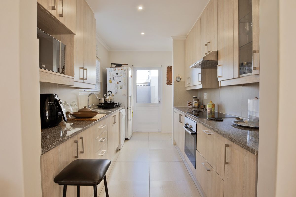 A well-maintained galley-style kitchen featuring light wood cabinetry, granite countertops, integrated appliances, and tiled flooring with direct access to the exterior.