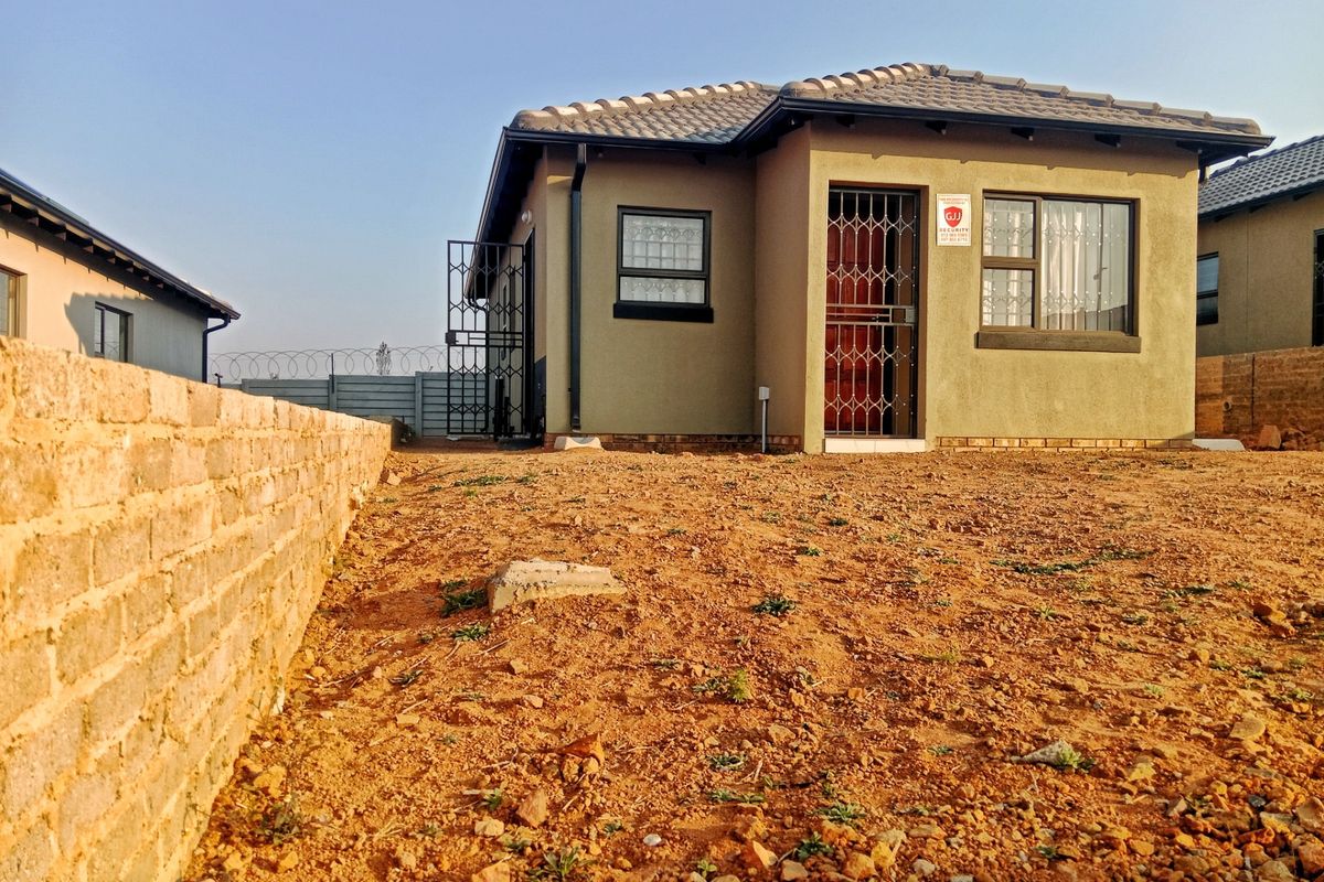 Rear view of a modern single-story house featuring a tiled roof, security gates, and a large, unlandscaped yard enclosed by a brick boundary wall.