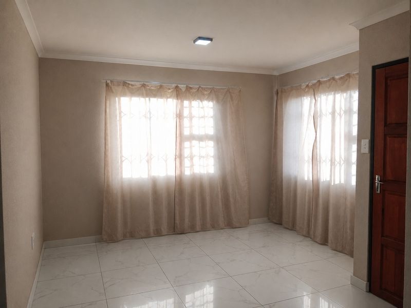 A bright, empty living room featuring neutral walls, white tiled flooring, and windows fitted with security bars and curtains.