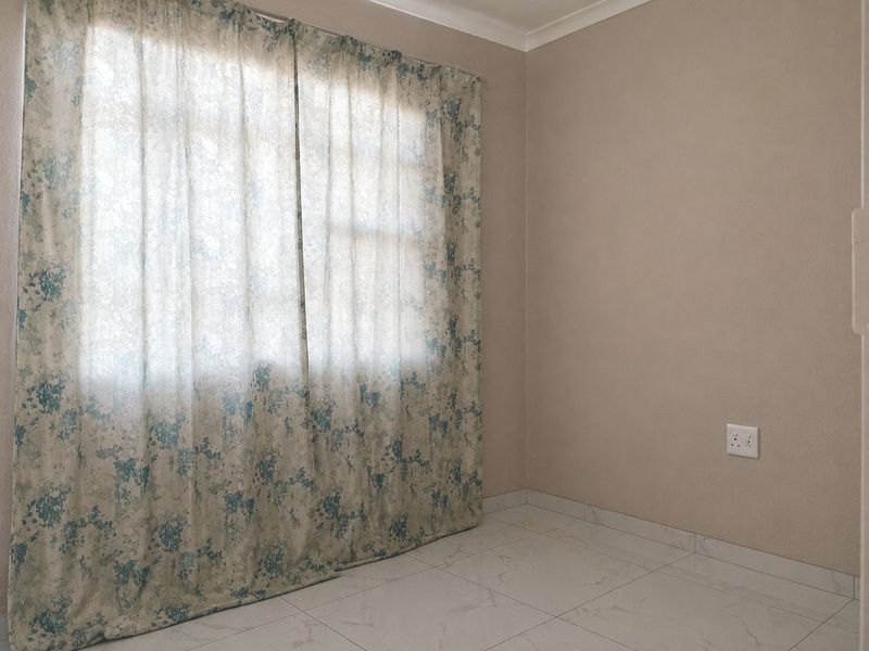 A clean, empty bedroom featuring neutral wall tones, tiled flooring, and a large window with curtains, providing a versatile space ready for personalization.