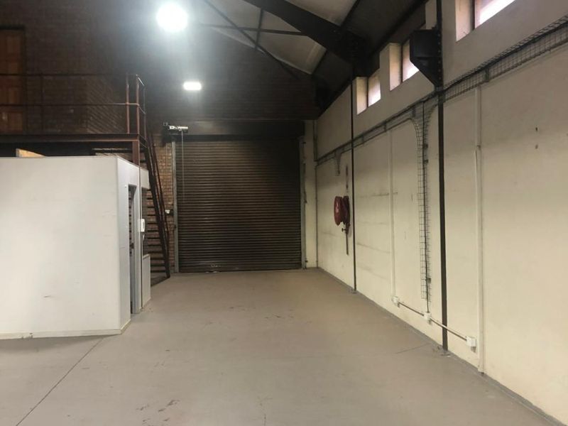 Spacious industrial utility area featuring high ceilings, exposed steel beams, a large roller shutter door, and a mezzanine level for additional storage or office space.