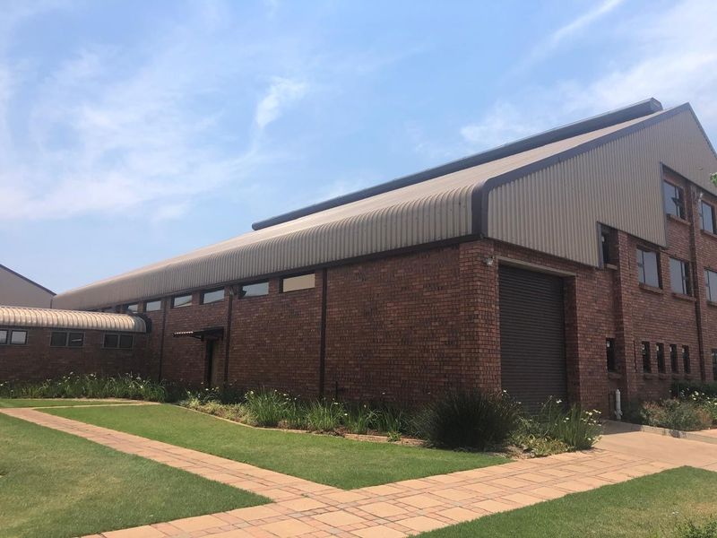 Exterior view of an industrial building featuring a brick facade, metal roofing, a large roller shutter door, and well-maintained surrounding paved walkways and gardens.
