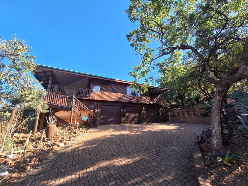 A two-story timber-clad house featuring a double garage, a paved driveway, and an elevated wooden balcony set within a landscaped garden.
