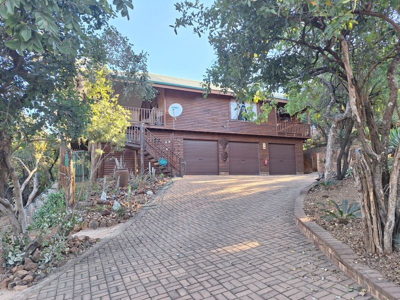 A charming double-story timber-clad home featuring a triple garage, paved driveway, and an elevated balcony nestled in a natural, wooded setting.
