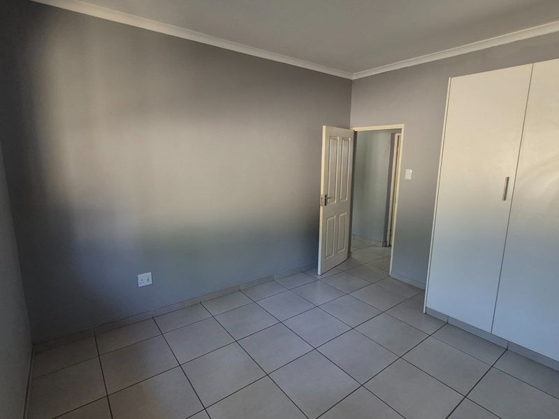 A clean, empty bedroom featuring neutral grey walls, tiled flooring, and white built-in cupboards with security bars on the window.