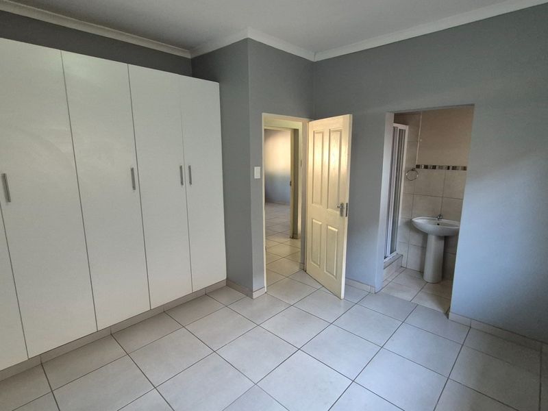 A clean, empty bedroom featuring built-in cupboards, neutral tiled flooring, and direct access to an en-suite bathroom with a pedestal basin and shower.