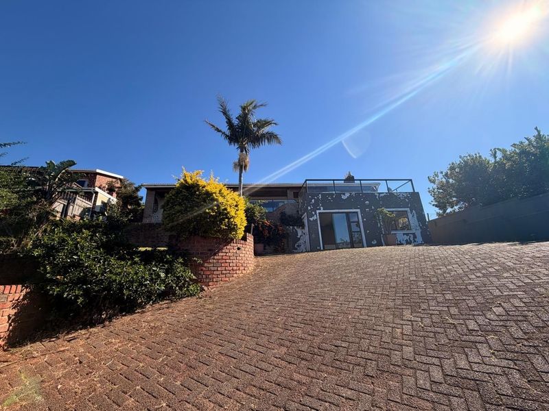 Front exterior view of the property featuring a brick paved driveway, retaining wall, and an upper-level balcony with metal railings.