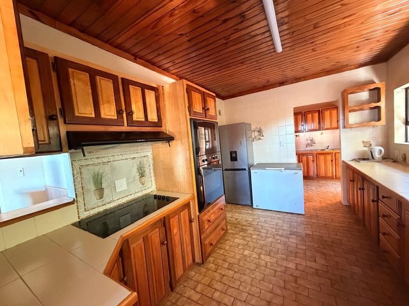 A spacious kitchen featuring extensive wooden cabinetry, tiled countertops, built-in oven, and a distinctive timber-clad ceiling, offering a functional layout with a separate scullery area.