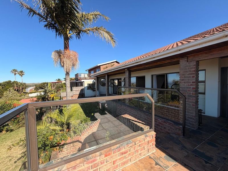 A spacious outdoor patio featuring brick pillars, glass balustrades, and tiled flooring, overlooking a landscaped garden area.