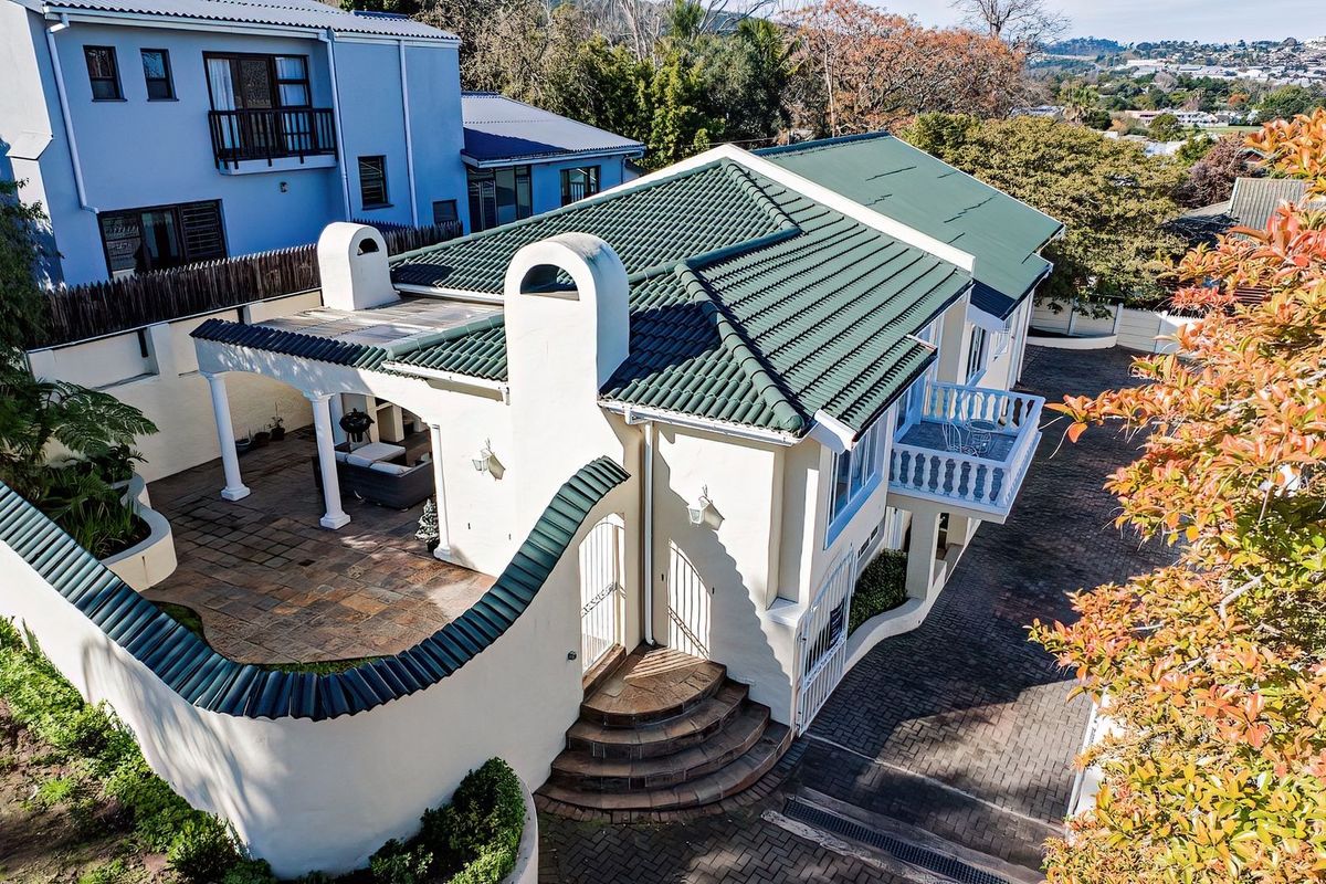 Aerial view of the property showcasing the tiled roof, paved patio area, balcony, and surrounding paved driveway with boundary wall.