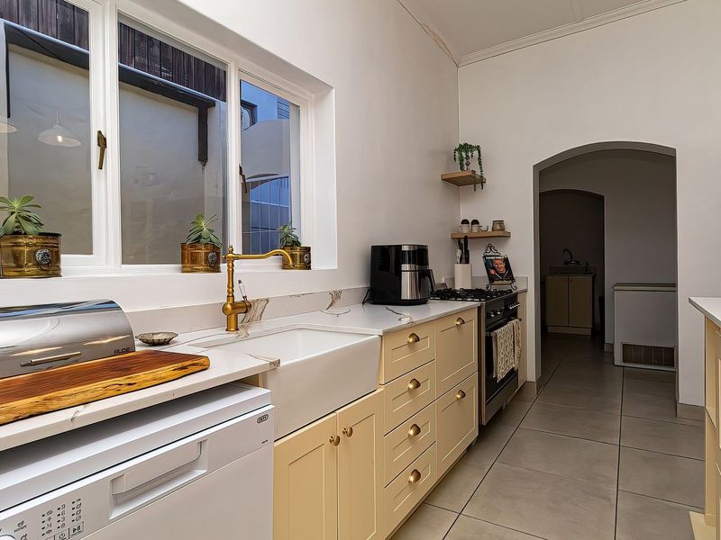 A well-maintained kitchen featuring shaker-style cabinetry, stone countertops, a farmhouse sink with brass fixtures, and tiled flooring, leading into a secondary utility area.