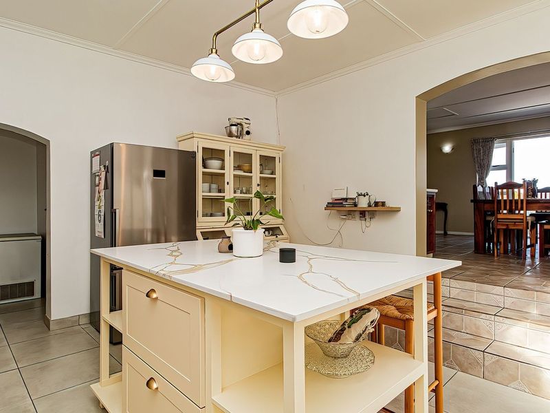 A bright kitchen featuring a central island with a stone countertop, pendant lighting, and tiled flooring, leading into an adjacent dining area.