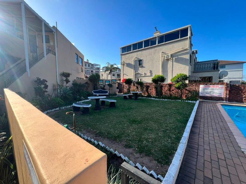 Communal outdoor area featuring a swimming pool, paved walkway, and a landscaped lawn with boundary walling in a residential apartment complex.