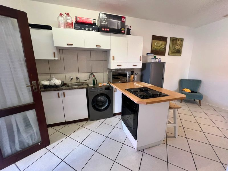 Compact kitchen featuring white cabinetry, tiled backsplash, built-in oven, hob, and integrated laundry plumbing, finished with neutral floor tiles.