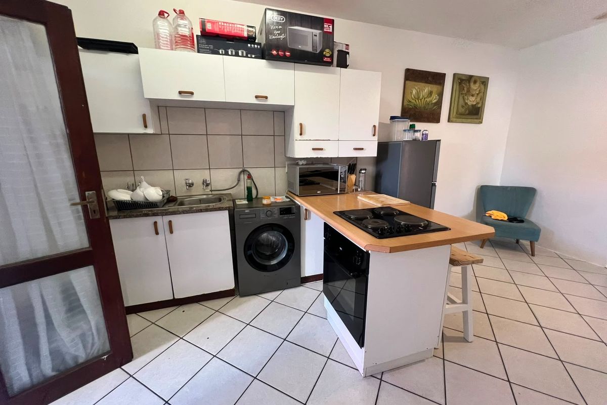 Compact kitchen featuring white cabinetry, tiled backsplash, built-in oven, hob, and integrated laundry plumbing, finished with neutral floor tiles.