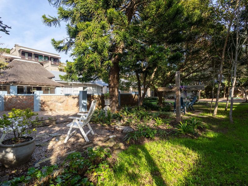A lush, mature garden area featuring established trees, gravel pathways, and a view of the property's brick boundary wall and thatched roof section.