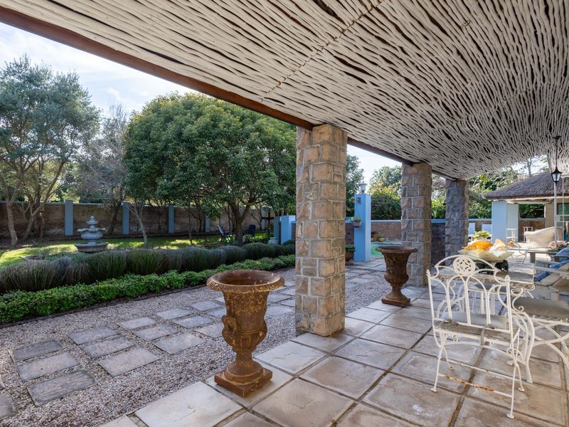 A spacious covered patio featuring rustic stone pillars and a thatched roof, overlooking a landscaped garden with a stone boundary wall.