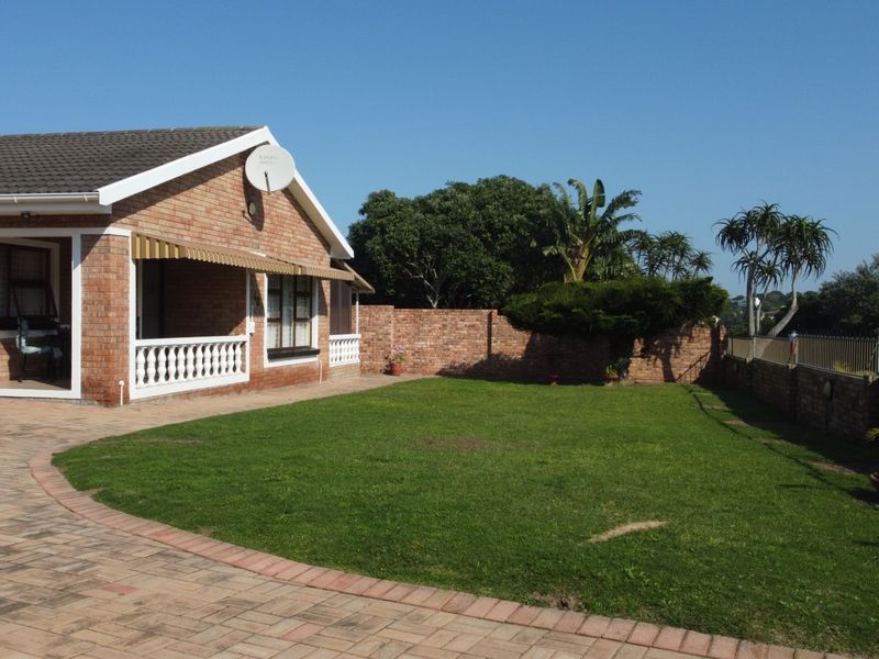 A well-maintained rear exterior view of a brick house featuring a paved driveway, a covered patio area with balustrades, and a spacious lawn.