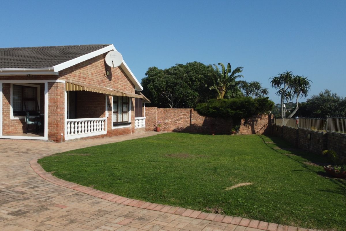 A well-maintained rear exterior view of a brick house featuring a paved driveway, a covered patio area with balustrades, and a spacious lawn.