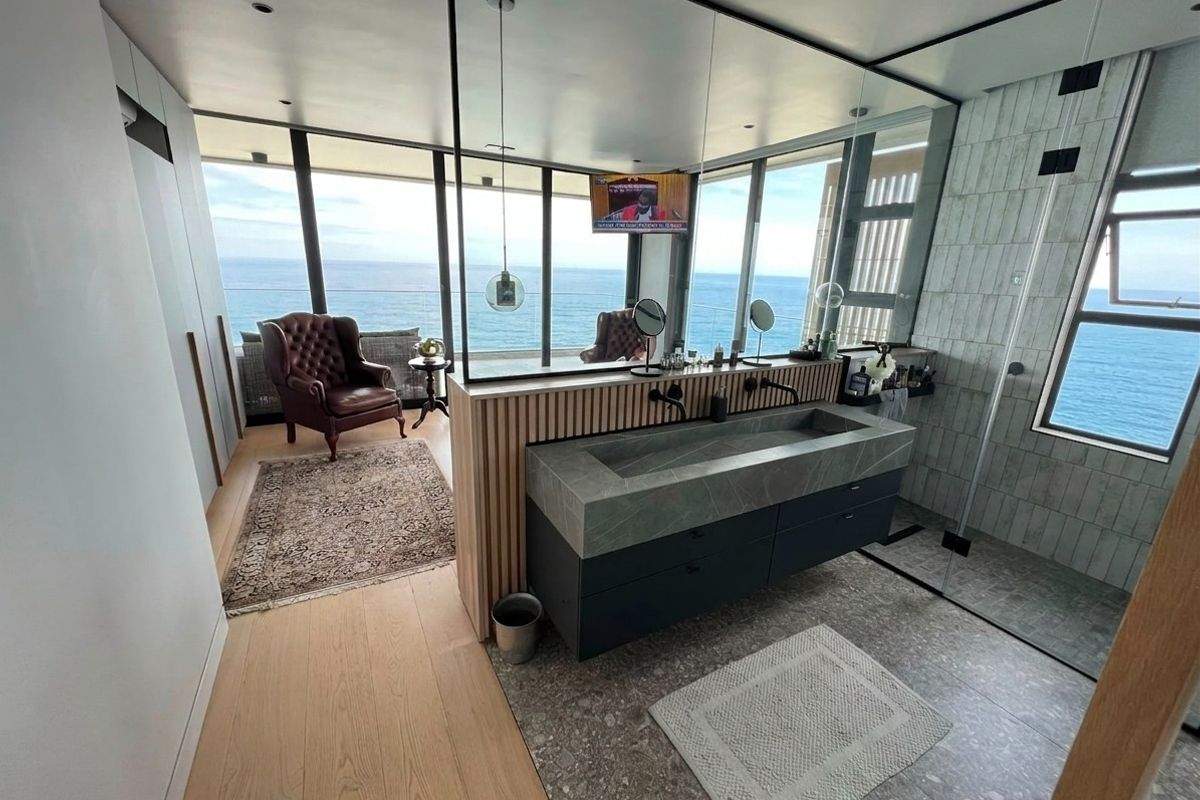Modern luxury bathroom featuring a stone double vanity, glass-enclosed shower, and floor-to-ceiling windows with panoramic ocean views.