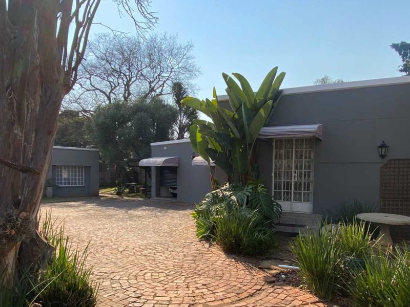 A well-maintained front exterior view featuring a cobblestone driveway, grey painted facade, and established garden landscaping.