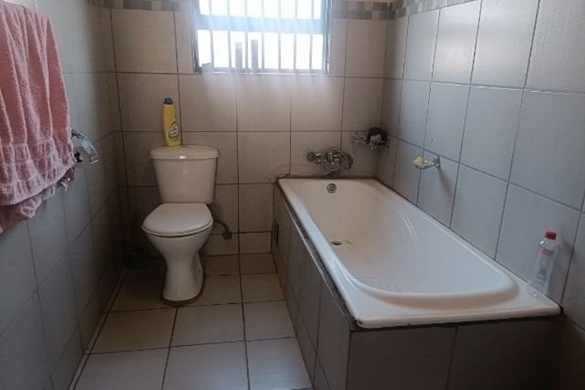 A functional bathroom featuring a standard bathtub, toilet, and full-height wall tiling with security bars on the window.
