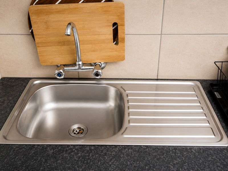 A clean kitchen sink area featuring a stainless steel basin, chrome faucet, and neutral ceramic wall tiling.