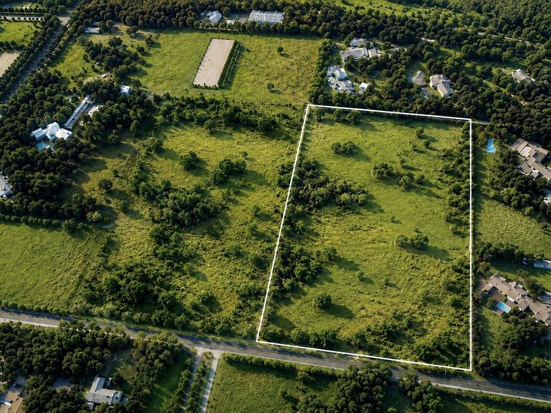 An aerial view of a large, rectangular vacant residential plot featuring established trees and open grassland, situated within a developed neighborhood.