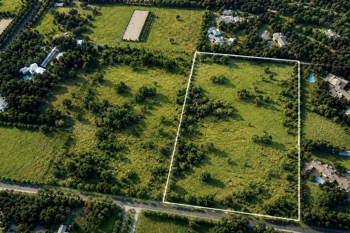 An aerial view of a large, rectangular vacant residential plot featuring established trees and open grassland, situated within a developed neighborhood.