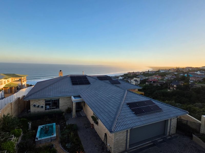 Aerial view of a coastal property featuring a tiled roof with solar panels, brick exterior, paved driveway, and a landscaped garden with a water feature.