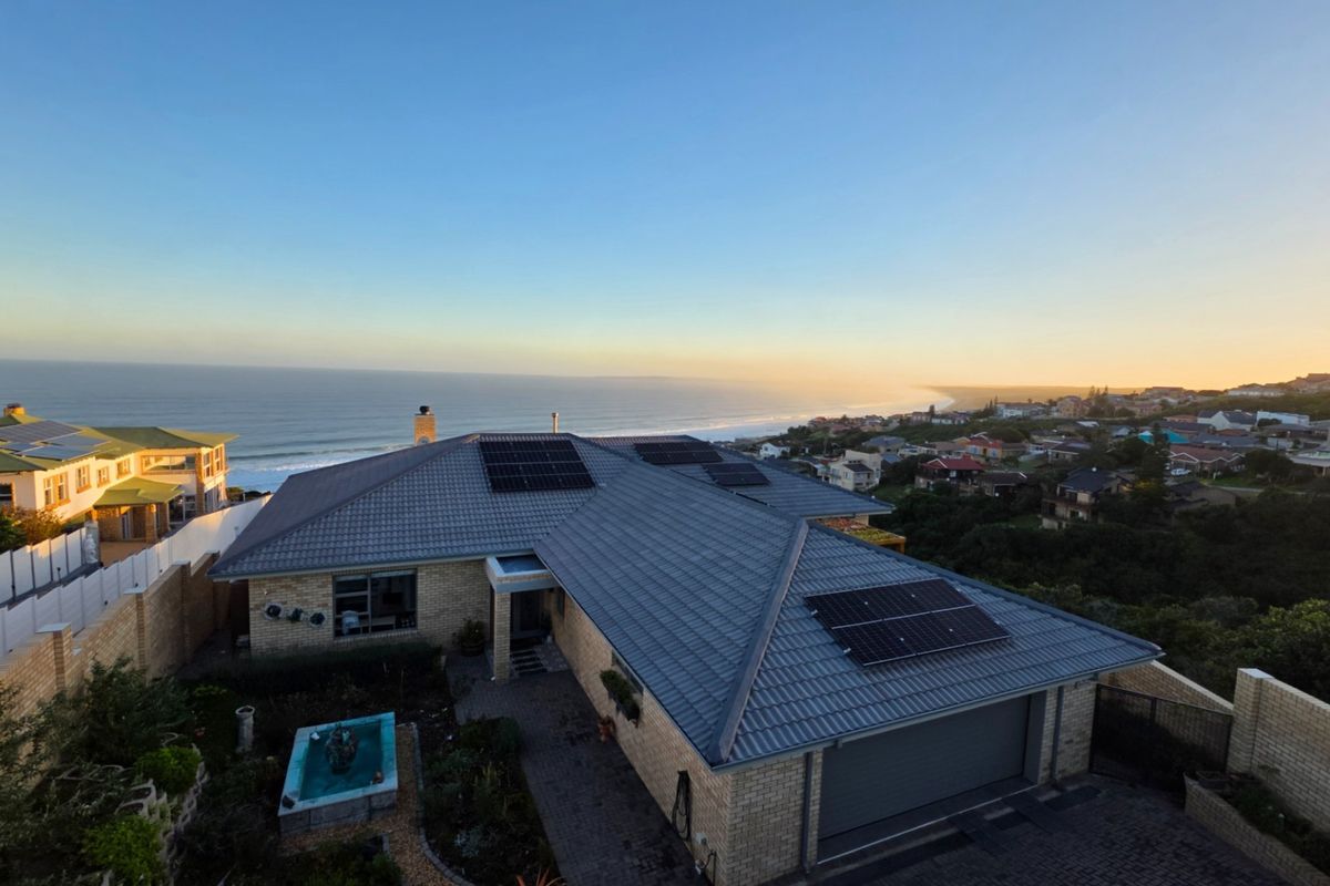 Aerial view of a coastal property featuring a tiled roof with solar panels, brick exterior, paved driveway, and a landscaped garden with a water feature.