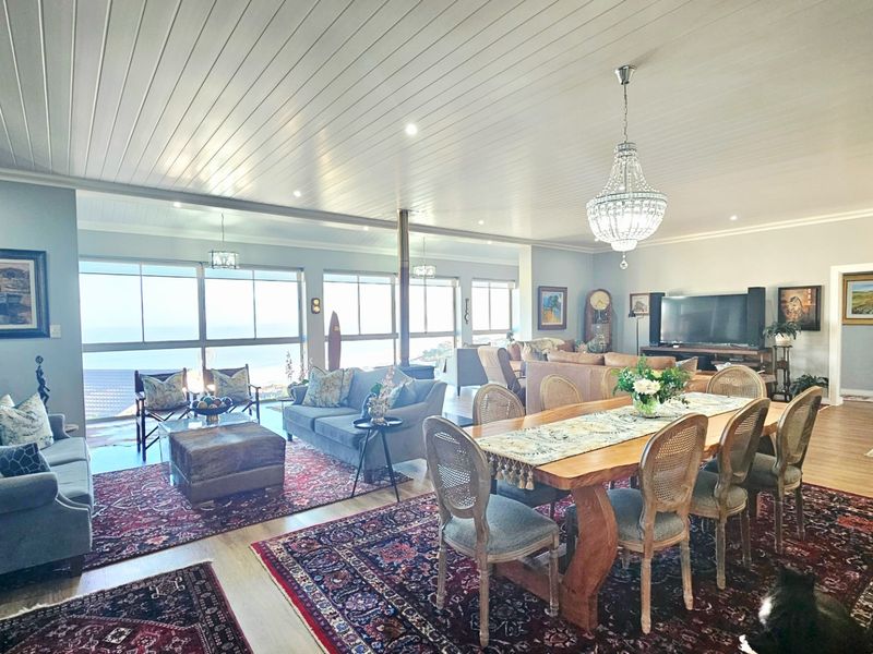 Spacious open-plan living and dining area featuring a wood-burning fireplace, tongue and groove ceiling, and expansive windows with panoramic coastal views.