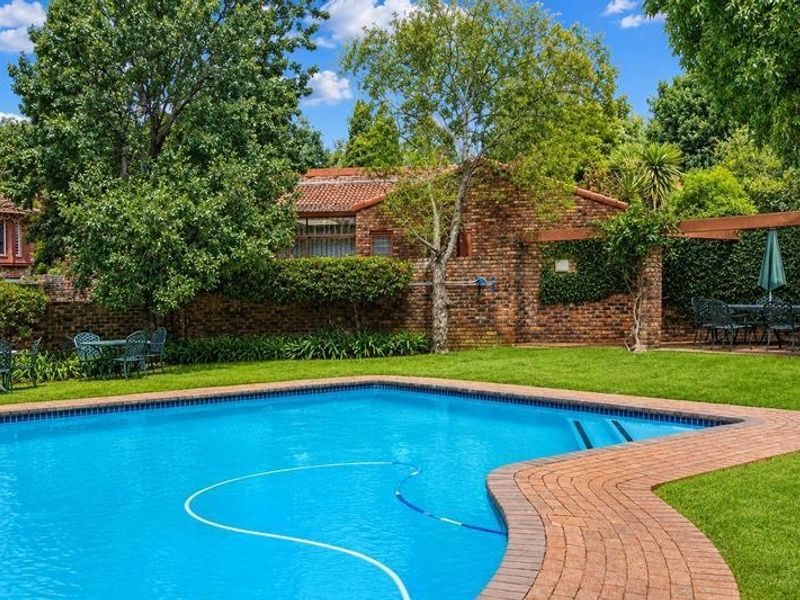 A well-maintained outdoor pool area featuring a swimming pool, brick paving, and a timber pergola set within a landscaped garden.