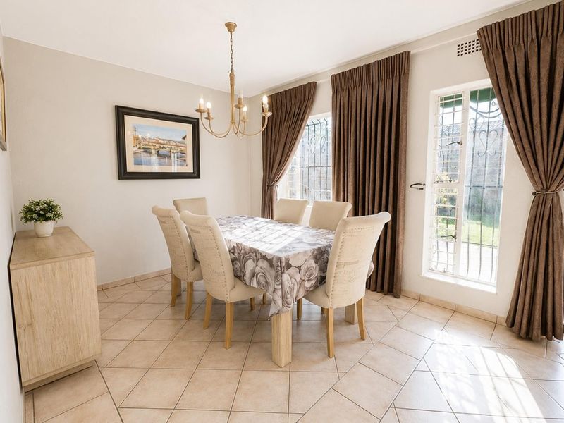 A bright dining room featuring ceramic tile flooring, a central chandelier, and large windows with security gates, offering direct access to the garden area.