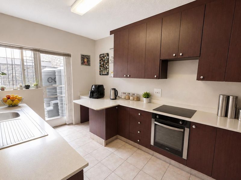 A functional kitchen featuring dark wood cabinetry, light countertops, an integrated electric hob and oven, and tiled flooring with direct access to an exterior area.